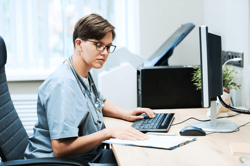 Concentrated young nurse in eyeglasses sitting at desk and using computer and papers while filling medical report